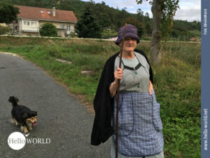 Begegnung auf dem Camino Portugues Dieses Bild vom Camino Portugues zeigt eine mit Schürze und Mütze bekleidete alte Frau, einen langen Stock in der Hand und hinter ihr ein schwarz-brauner Hund.