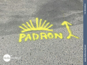 Das Bild von der letzten Camino Portugues Etappe zeigt eine grau asphaltierte Stra&szlig;e, auf der mit gelber Farbe ein Wegweiser nach Padron aufgemalt ist.