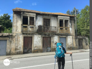 Hier sieht am eine Pilgerin auf dem Camino Portugues K&uuml;stenweg, vor ihr die Stra&szlig;e, dahinter ein altes, zerfallenes Haus.