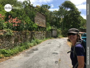 Dorfatmosphäre an der Costa da Morte Das Bild von der Costa da Morte zeigt eine kleine Dorfstraße links von einer steinernen Mauer mit viel grün und roten Blumen umgeben, im Vordergrund eine Wanderin mit Rucksack und Schildmütze.