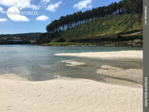 Die Ria de Lires an der spanischen Nordküste Das Bild von der spanischen Nordküste zeigt die Ria de Lires Richtung Landesinnere, im Vordergrund Sand und Wasser, dahinter Häuser, rechts eine Baumreihe.