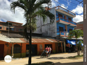 Diese Aufnahme aus der Serie Bilder aus S&uuml;damerika zeigt zwei Personen auf einem Motorrad, die an bunten H&auml;usern in einem Dorf vorbeifahren.