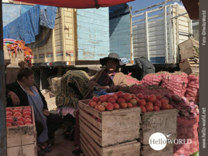 Zentnerweise Gemüse direkt vom Feld Das Bild, aufgenommen auf dem bolivianischen Mercado el Morro, zeigt zwei Marktfrauen vor Tomatenkisten, im Hintergrund Lastwagen.