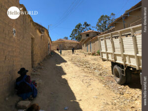 Das Bild von Tarabuco, Bolivien, zeigt eine unbefestigte, staubige Stra&szlig;e am Ortsrand, rechts parkt ein Transporter, links sitzt eine Frau, ganz hinten spielen Kinder.