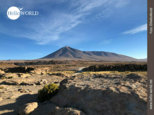 Dieses Bild aus Bolivien zeigt in der Ferne einen Vulkan und im Vordergrund die Ebene mit ihren braunen Felsen und einer Fahrbahn.