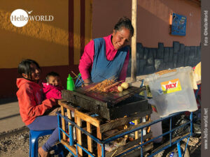 Das Bild aus Cusco, Peru, zeigt eine lachende Frau vor ihrem Grill, daneben sitzt ein M&auml;dchen und h&auml;lt ein Kleinkind auf dem Scho&szlig;.