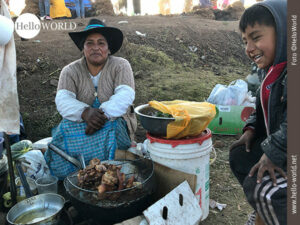 Dieses Bild aus Cusco, Peru, zeigt eine Frau mit Hut und ein lachendes Kind, die vor einem Topf mit fritierten Fleischst&uuml;cken sitzen.