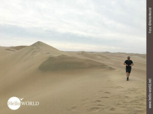 Spuren hinterlassen: Dünenwanderung in Peru Dieses Bild zeigt einen Mann in kurzen Hosen und mit Kappe bekleidet, wie er an einer Düne in der Wüste entlang läuft.